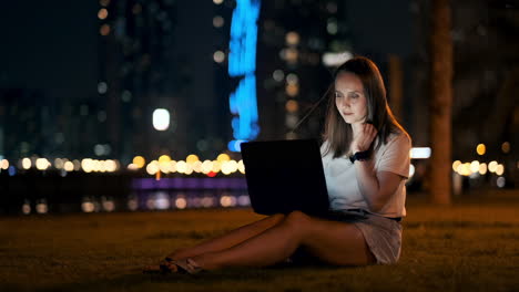 A-young-female-student-with-a-laptop-at-night-in-the-city-looking-at-the-computer-screen-and-typing-with-his-hands-on-the-keyboard.-Remote-work-on-the-Internet.-The-student-does-the-work.-Night-life-in-the-city
